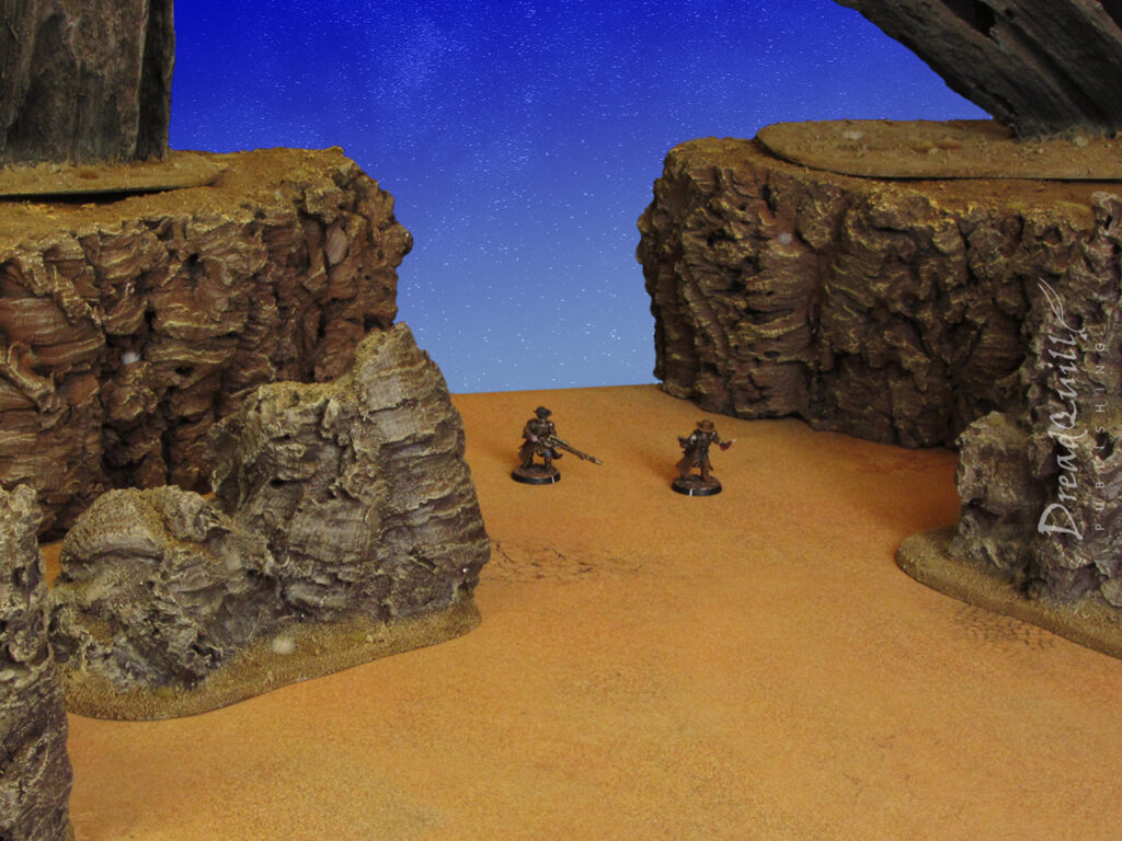 An ash wastes scene with modular hills in the background, some rocky outcrops in the foreground, a desert blue background and two necromunda fighters in the distant horizon