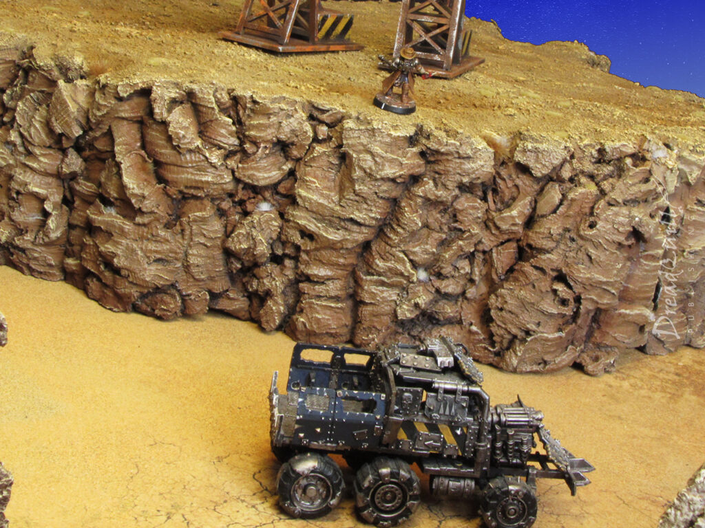 A necromunda genestealer cultist prepares to drop a stick of dynamite from atop some modular hills onto a ramshackle vehicle beneath him